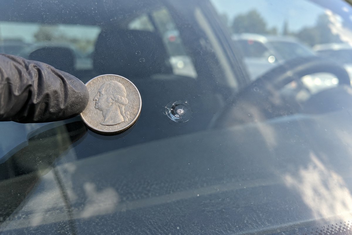 A US quarter held against a windshield beside a small round rock chip, smaller than the coin