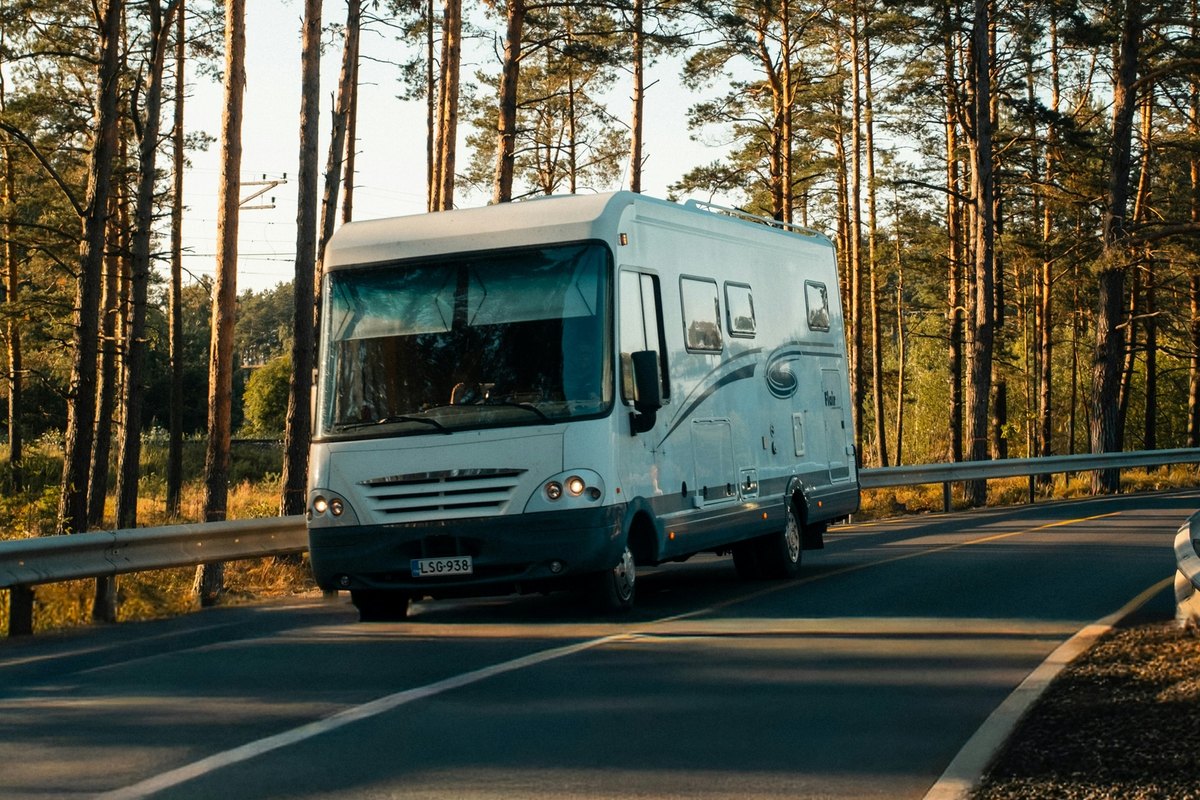 White Class A motorhome driving toward the camera on a two-lane road lined with tall pines