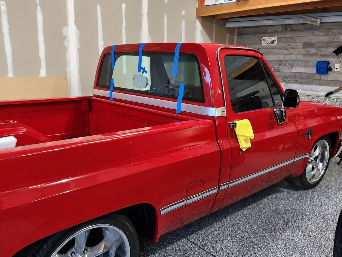 Red classic Chevrolet pickup with polished wheels in a garage, its newly fitted rear cab window held by blue tape