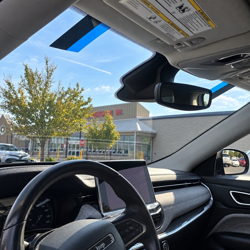 Driver's view through a newly fitted Jeep windshield, two strips of blue tape along its top edge and a parking lot beyond