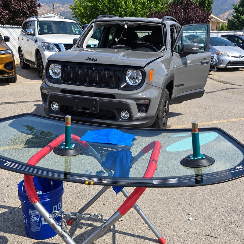 Gray Jeep Renegade in a parking lot with its windshield removed and new glass on a stand