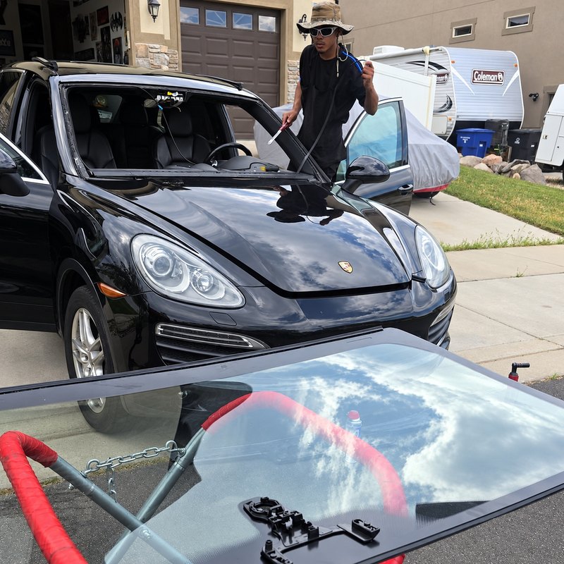 Technician in a bucket hat working along the empty windshield frame of a black Porsche Cayenne, the new glass on a stand